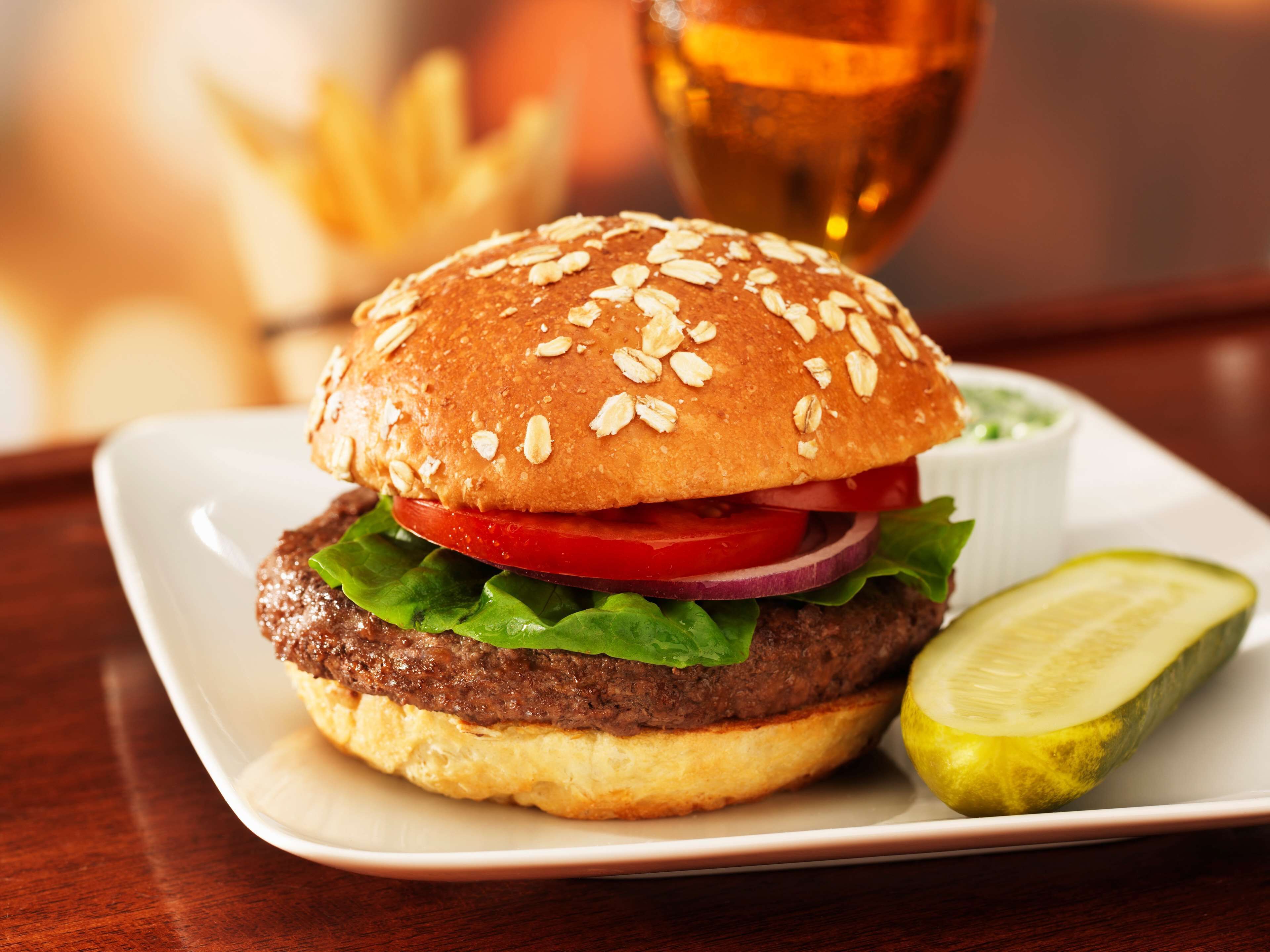 A hamburger with a sesame seed bun, lettuce, tomato, and a pickle on the side, served on a white plate.