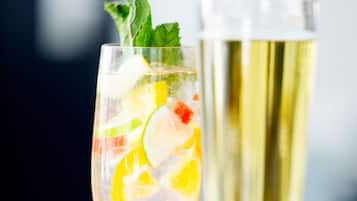 A hand holding a glass with a cocktail, garnished with lime and mint, and a tall glass of champagne in the background.