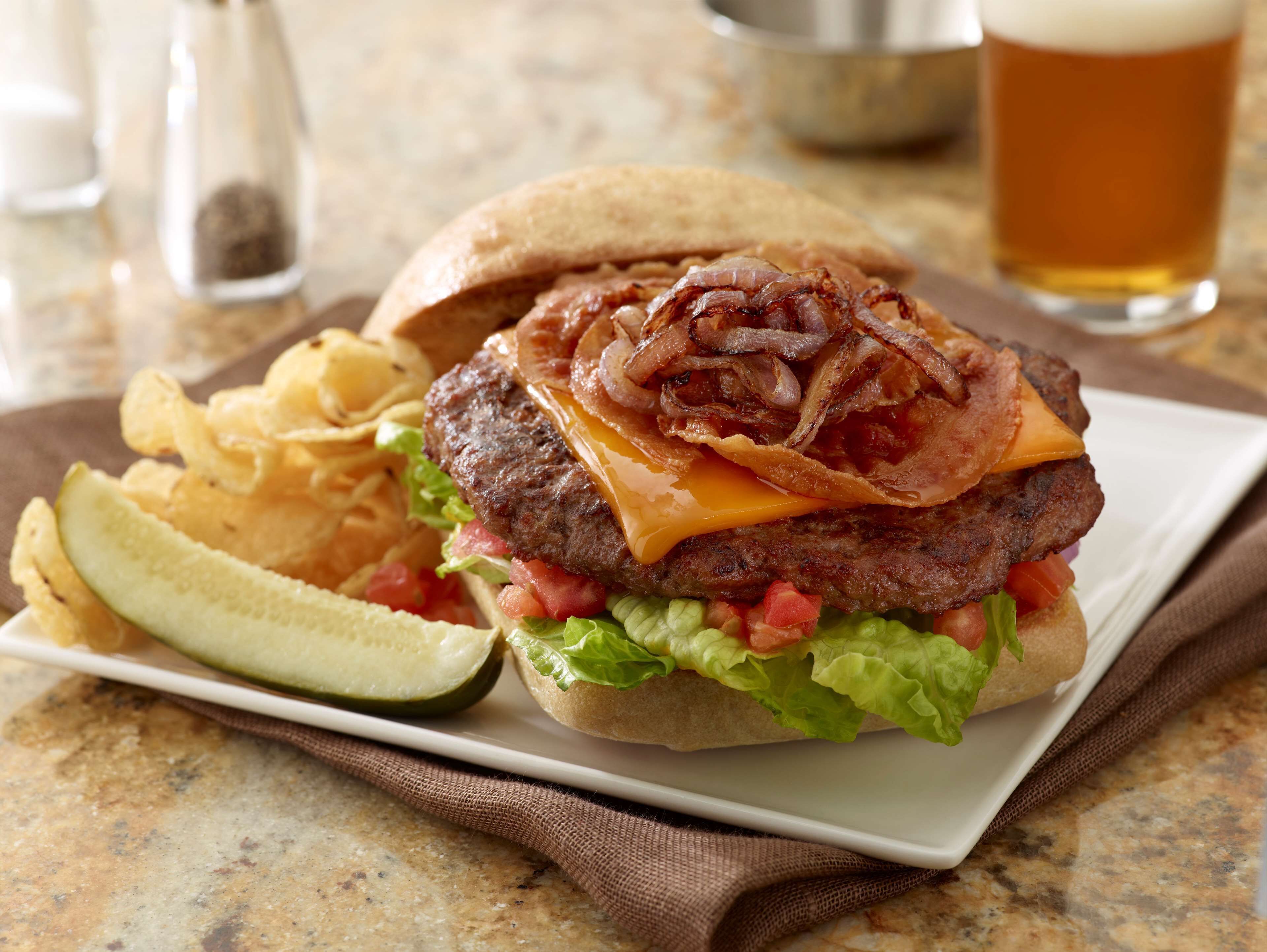 A burger with bacon, cheese, lettuce, and tomato, served with chips and a pickle on a plate.