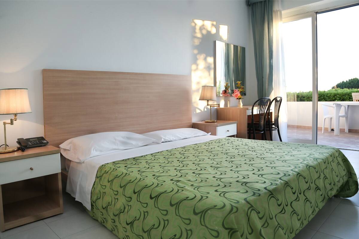 A hotel room with a bed, bedside lamps, a nightstand, a chair, a table, and a view of the outdoors.