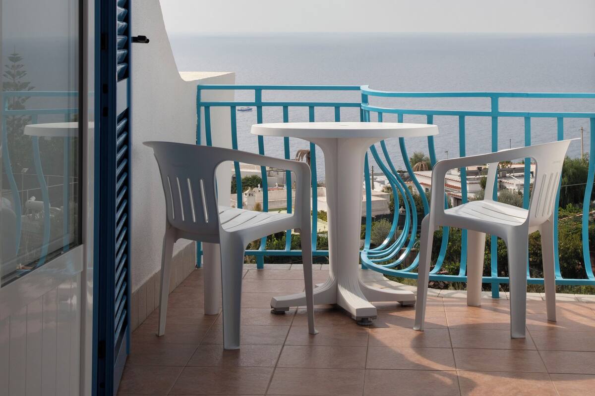 A balcony with white plastic chairs and a table, overlooking a coastal view.