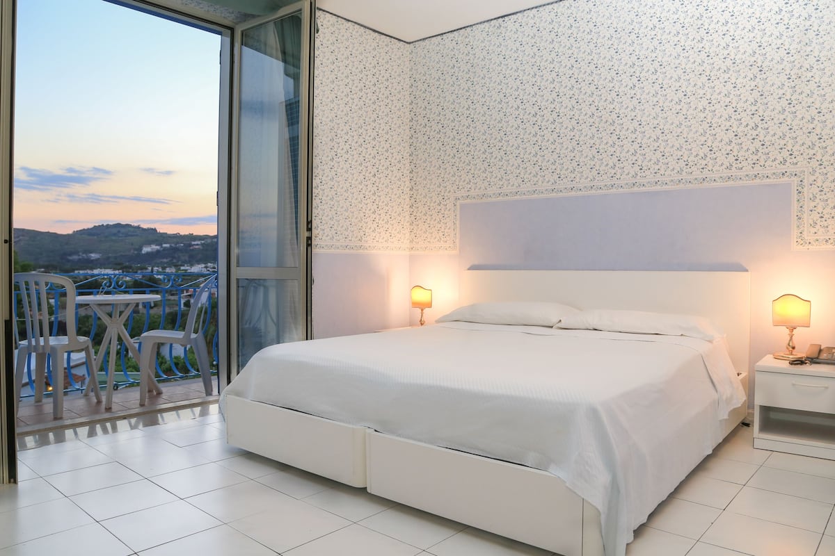 A bedroom with a large bed, two bedside lamps, a view of a balcony with chairs, and a patterned wall.