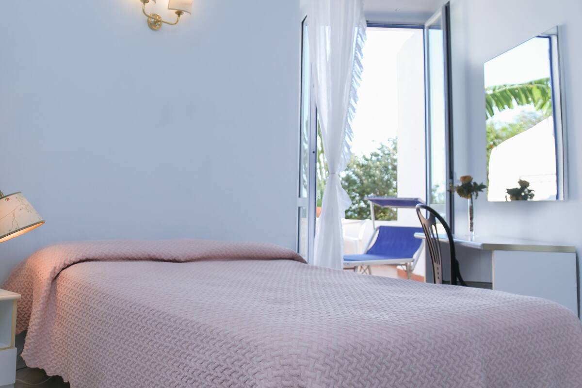 A bedroom with a bed, a nightstand, a chair, a window with curtains, and a wall-mounted light fixture.