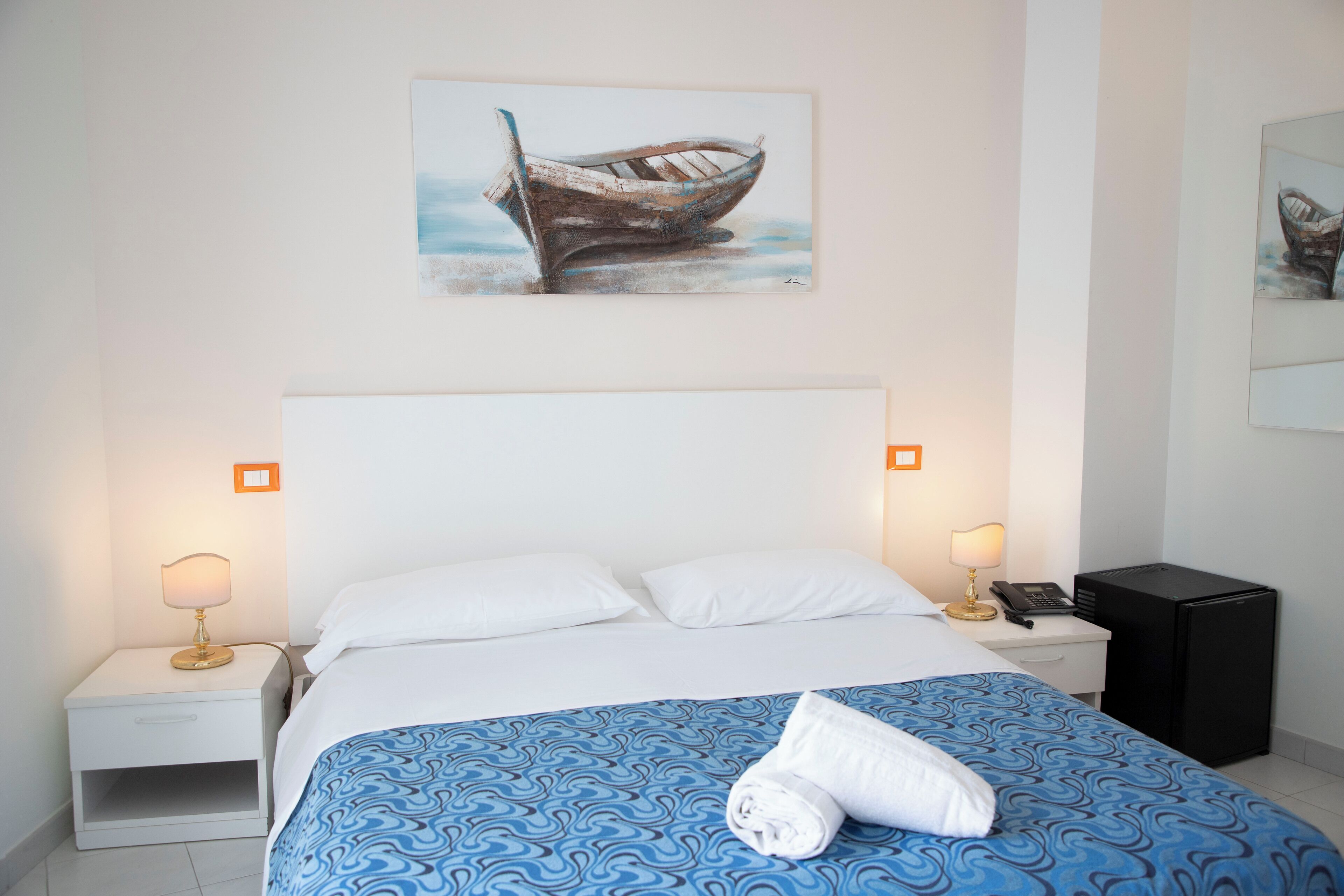 Une chambre d’hôtel avec un lit, deux tables de chevet, une petite table d’appoint, un petit réfrigérateur noir et un tableau représentant un bateau accroché au mur.