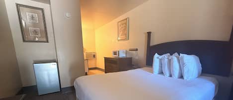 A hotel room with a bed, a desk, a chair, a small refrigerator, and a wall-mounted air conditioning unit.