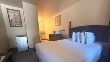 A hotel room with a bed, a desk, a chair, a small refrigerator, and a wall-mounted air conditioner.