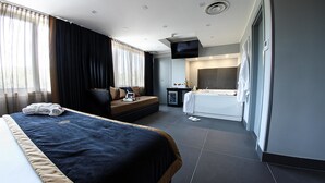 A modern hotel room with a large bed, a sofa, a small table with a bottle and glasses, a TV, and a bathroom with a bathtub and toilet. - Best Western JFK Hotel (Naples)