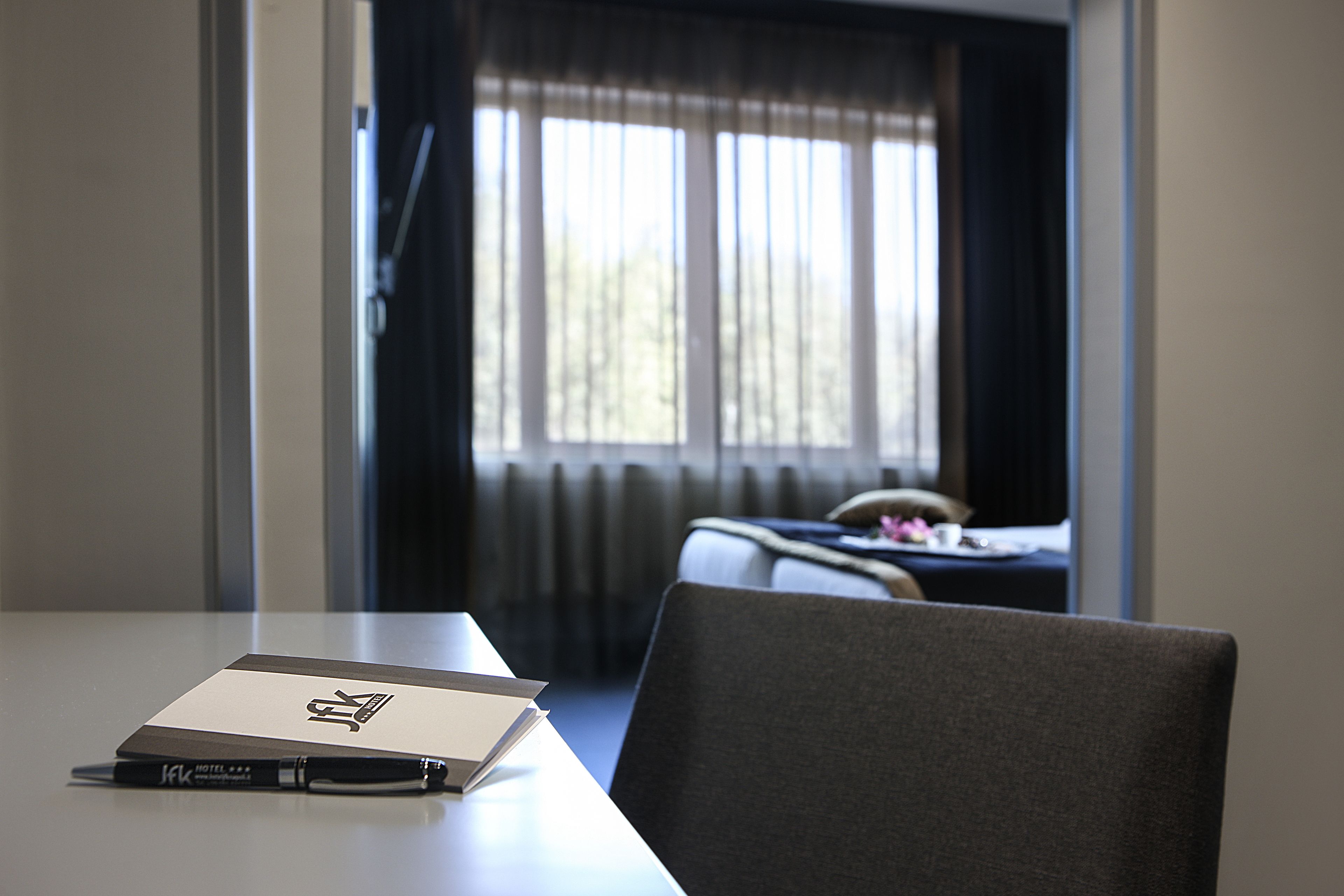 A hotel room with a bed, a window with curtains, a desk with a notebook and pen, and a chair.