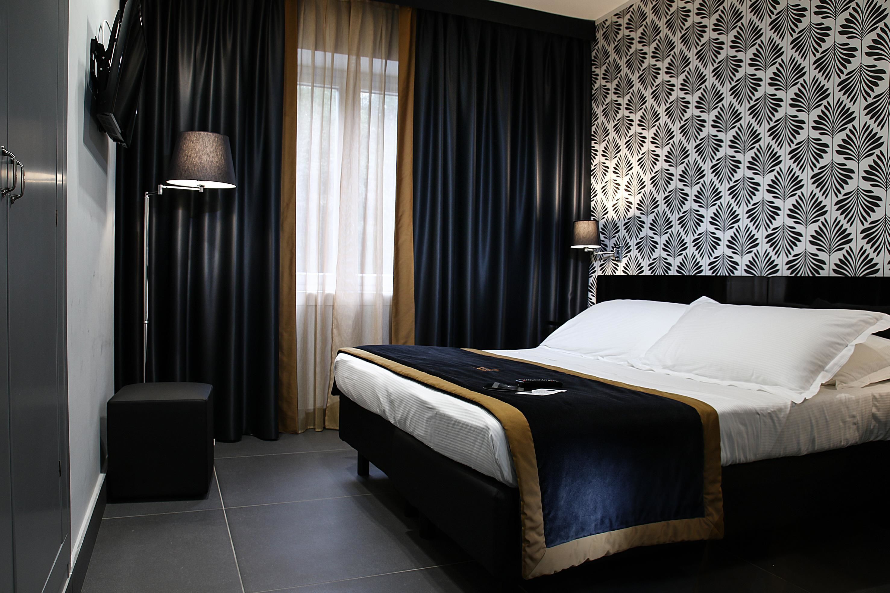 A modern hotel room with a large bed, a side table, a lamp, and a patterned wallpaper.