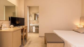 Habitación de hotel con cama, escritorio con televisor, baño con lavamanos y espejo, y una banca.