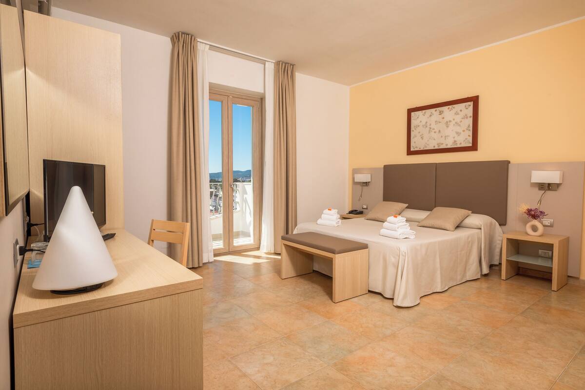 A hotel room with a bed, bedside tables, a desk with a lamp, a TV, and a balcony with curtains.