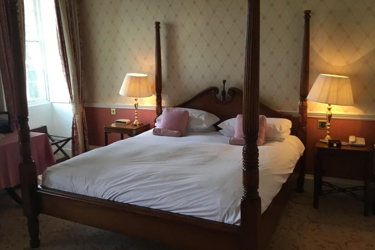 A four-poster bed with white linens, two bedside tables with lamps, and a window with curtains.
