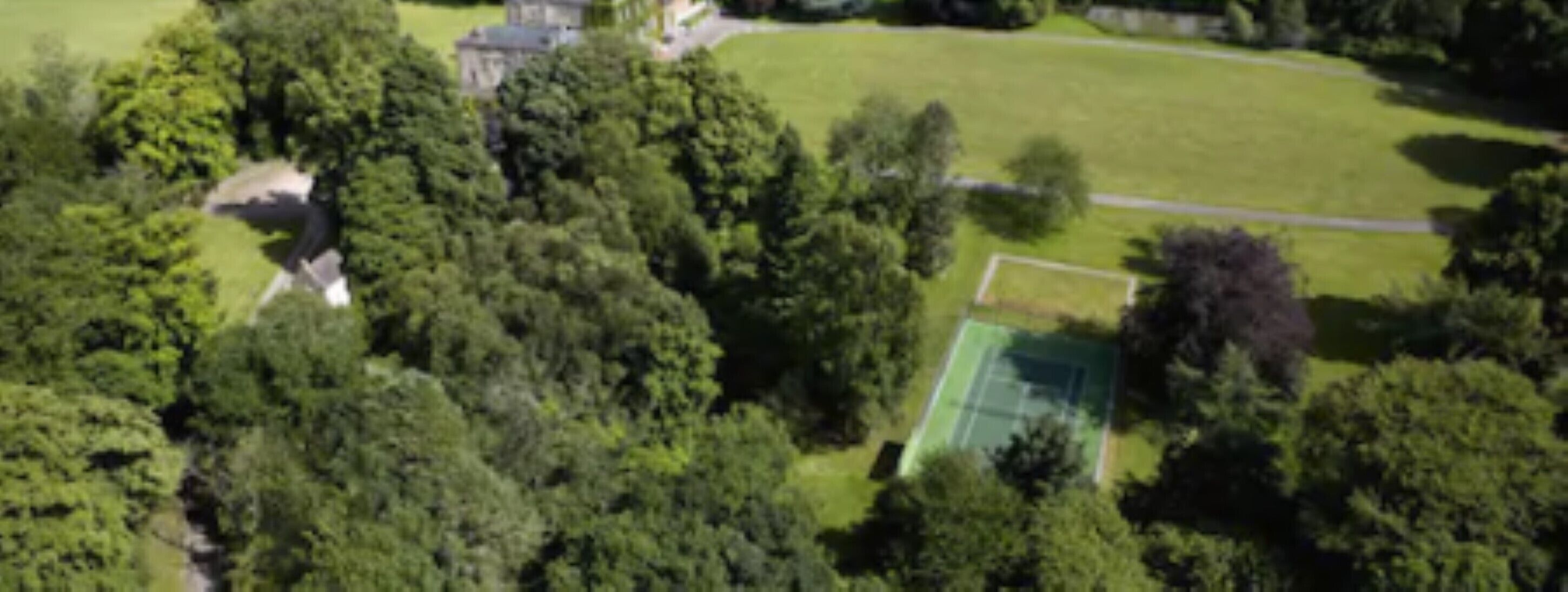 Vue aérienne d’une propriété dotée d’une végétation luxuriante, d’un court de tennis et d’un bâtiment à la façade en pierre.