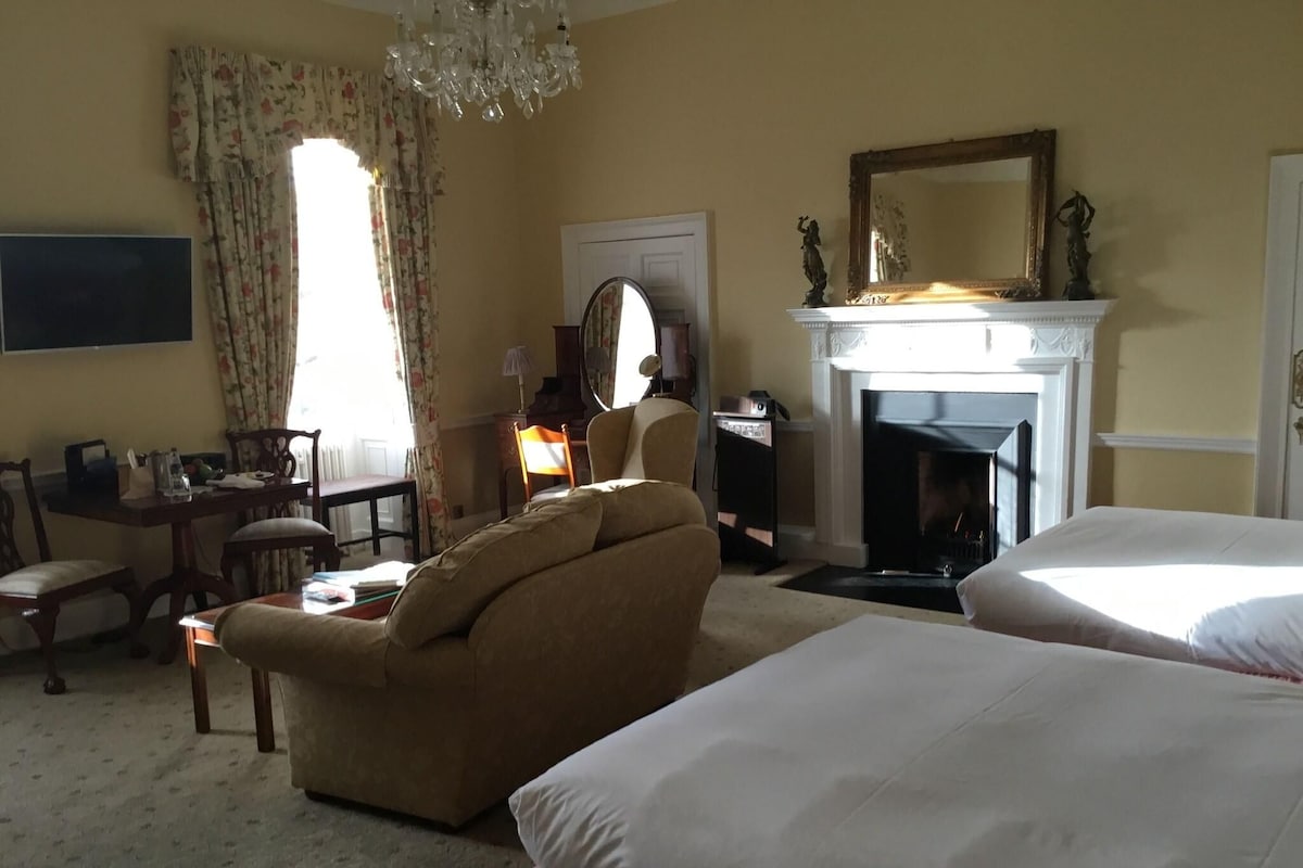 A hotel room with a sofa, two beds, a fireplace, a mirror, and a chandelier.
