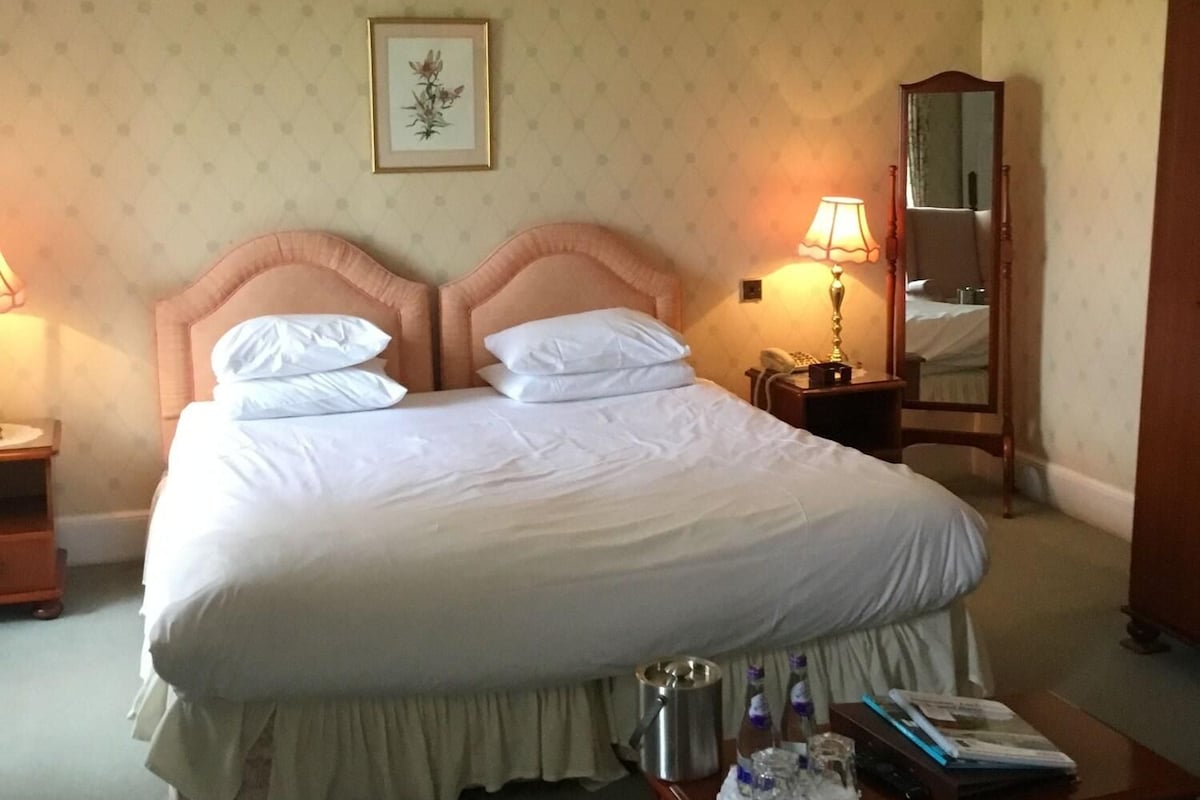 A neatly made bed with a headboard, a bedside table with a lamp, a wooden nightstand, a mirror, and a small table with a bottle and a book.