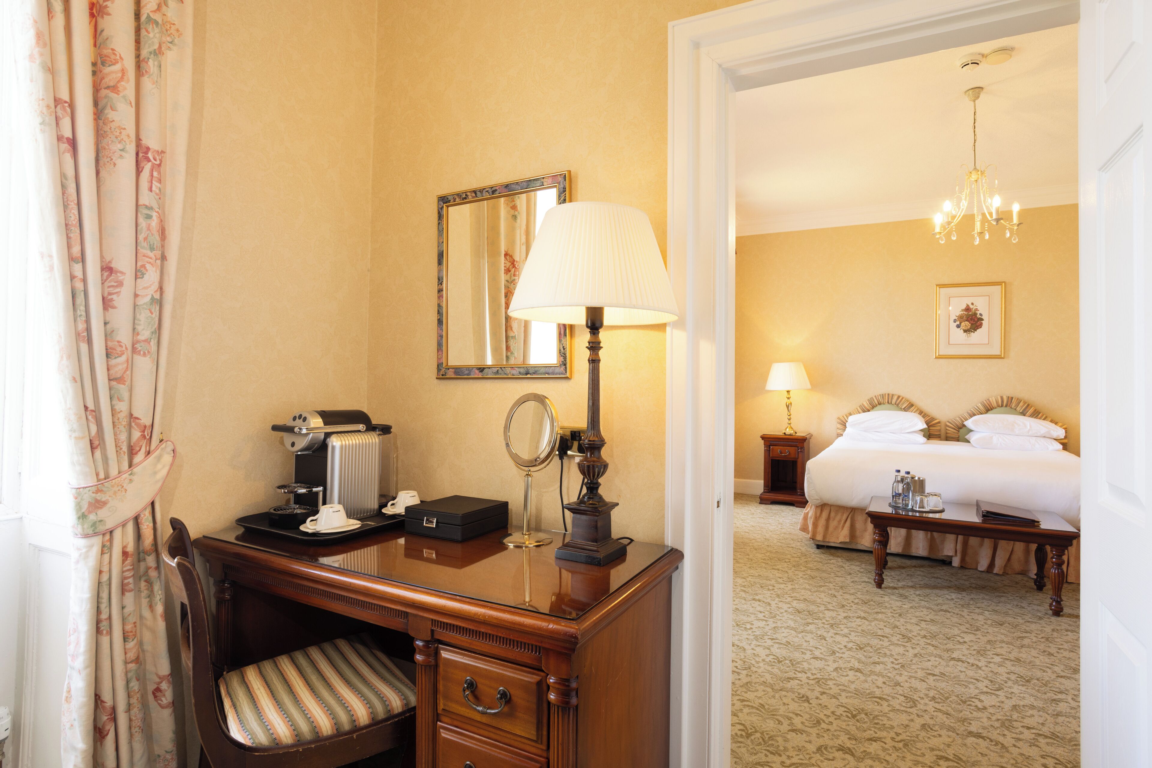 A hotel room with a wooden desk, a chair, a bed, a nightstand, and a chandelier.