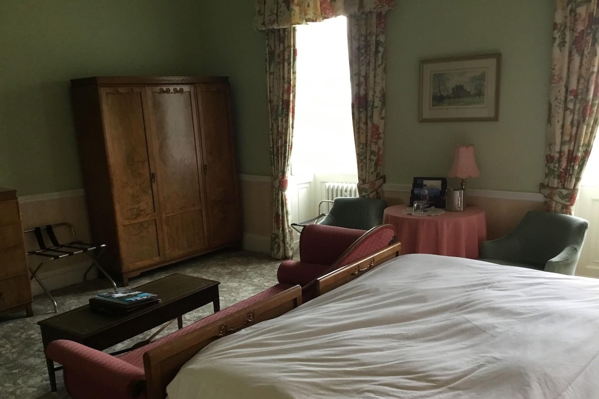 A hotel room with a large bed, a wooden wardrobe, a desk with a chair, a small round table with a chair, and a window with curtains.