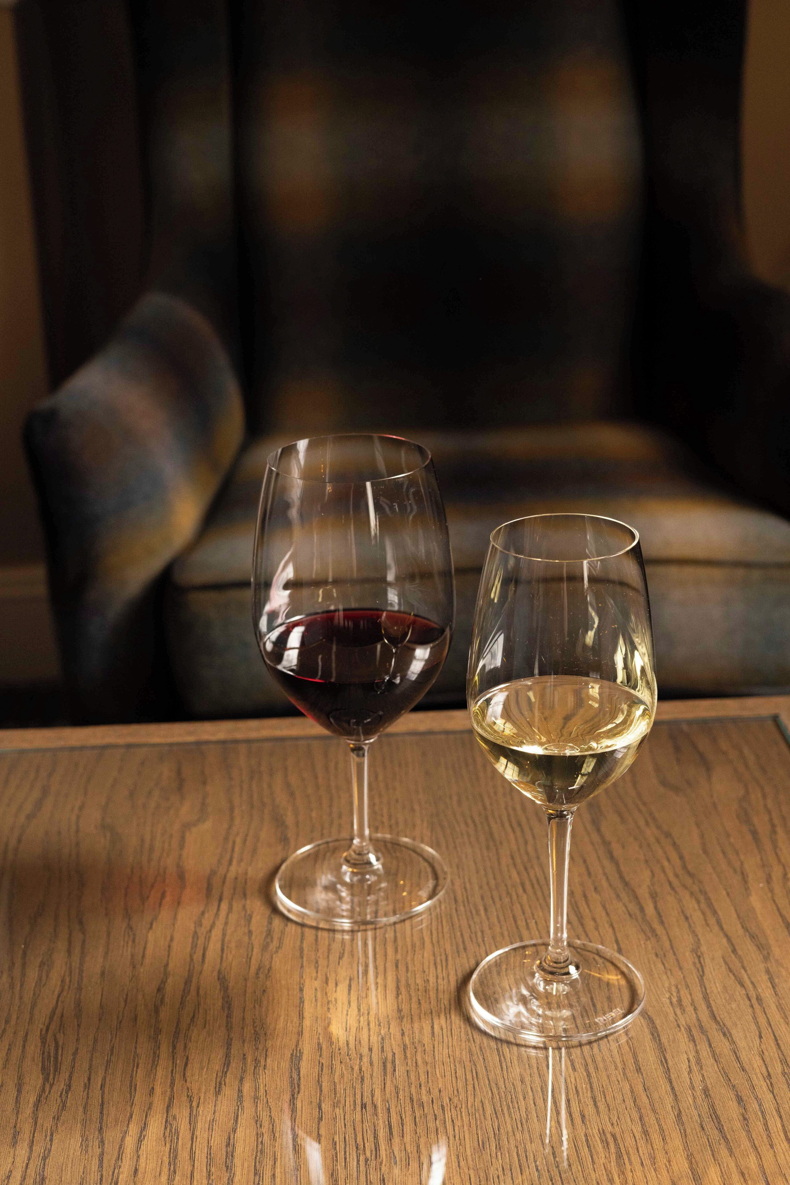 Two wine glasses on a wooden table, one with red wine and one with white wine, in a dimly lit room with a leather couch in the background.