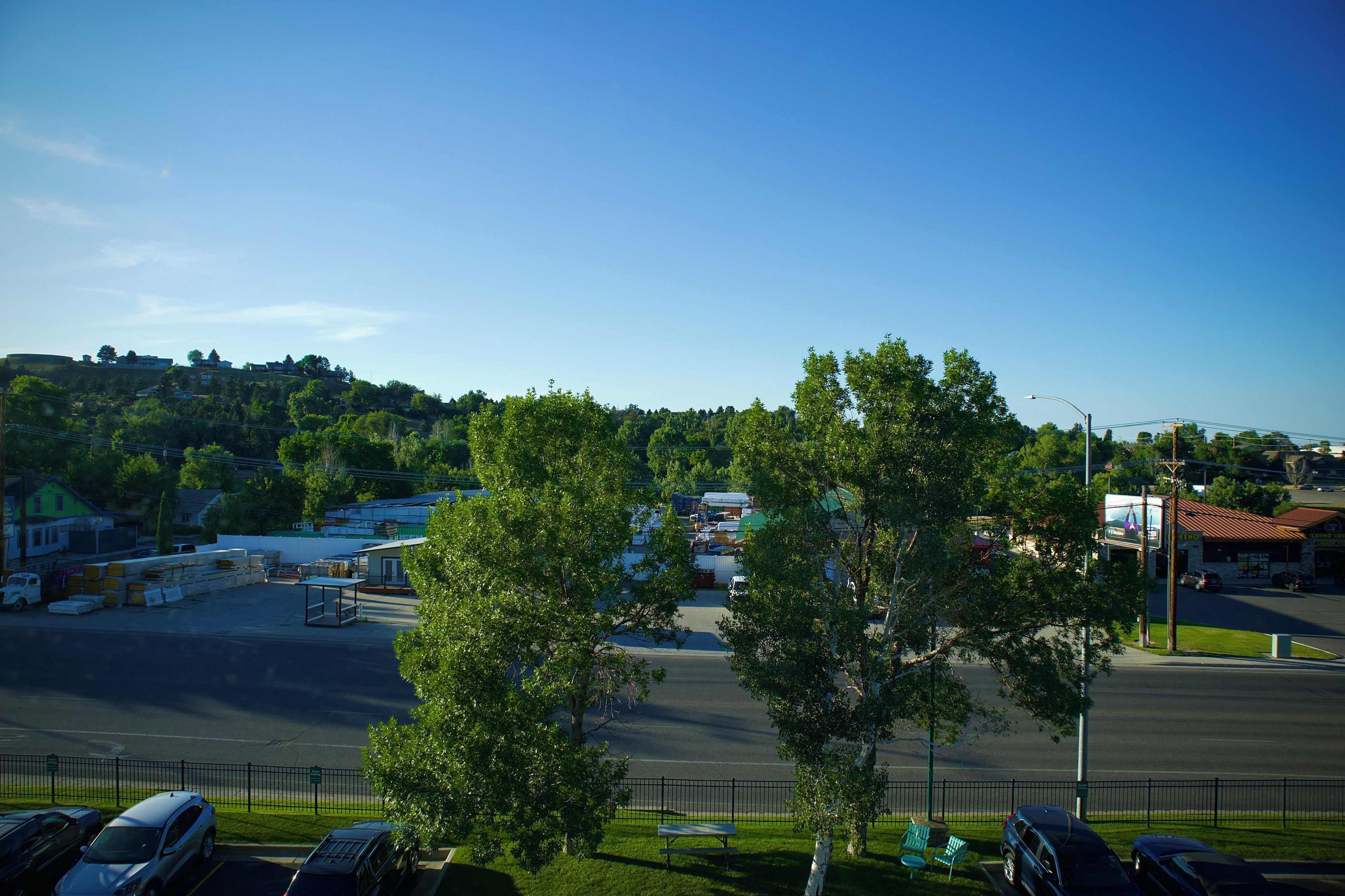 A parking lot with cars, a building with a green roof, and a hill with trees in the background.
