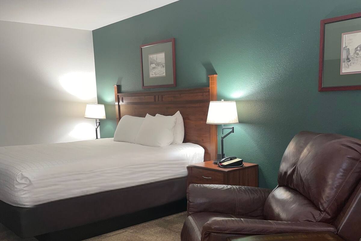 A hotel room with a bed, a leather armchair, a nightstand with a lamp, and two framed pictures on the wall.
