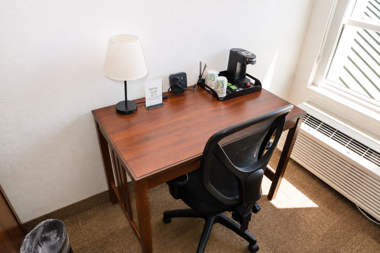 A hotel room with a wooden desk, a black office chair, a lamp, a coffee maker, and a sign with the hotel name.
