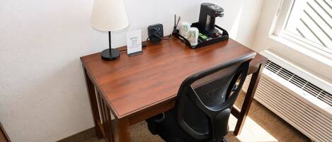A hotel room with a wooden desk, a black office chair, a lamp, a coffee maker, and a sign with the hotel name.