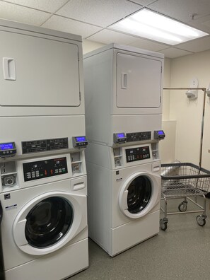 A laundry facility with stacked front-loading washers and dryers, control panels, and a cart. - Boothill Inn And Suites (Billings)