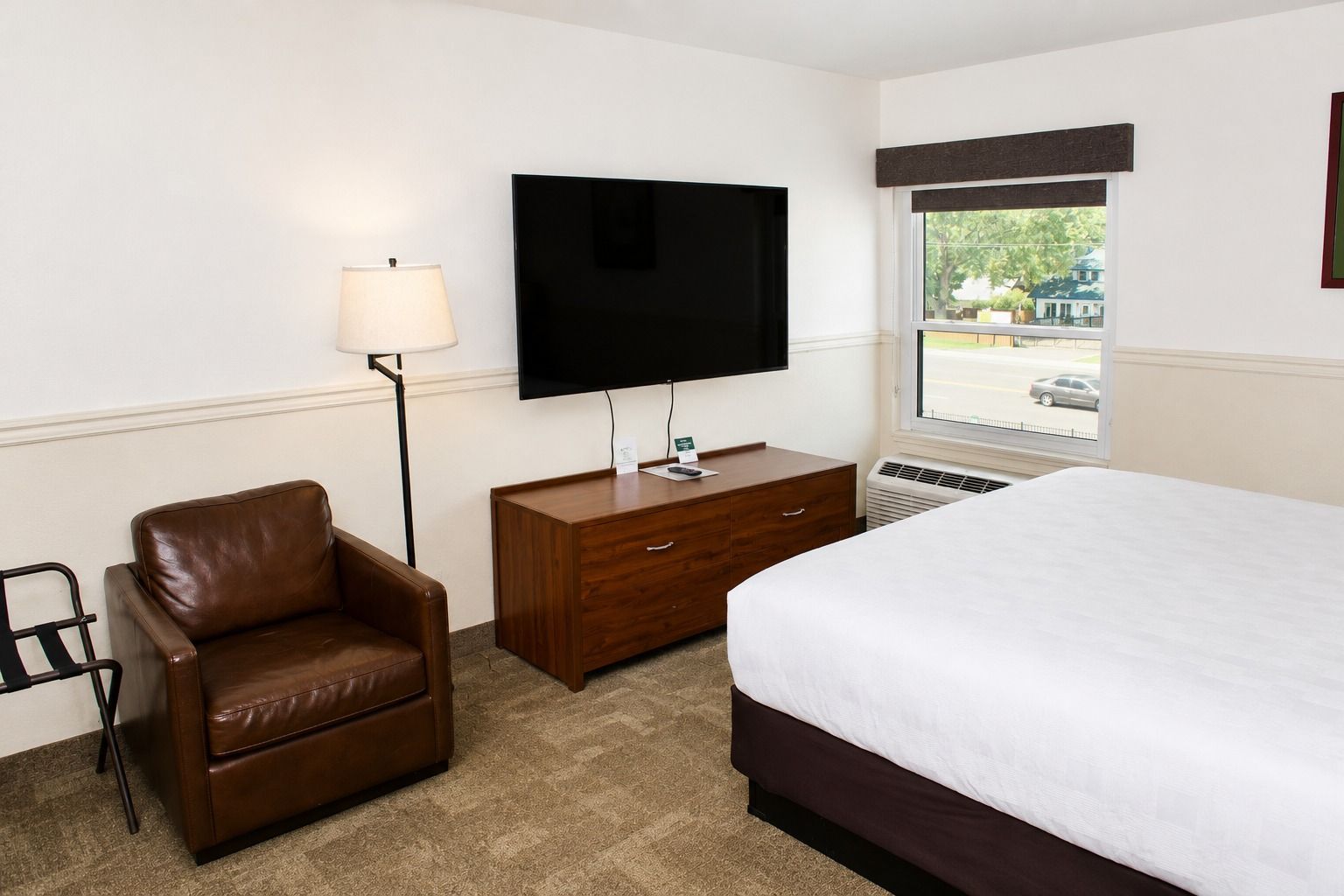 A hotel room with a bed, a desk with a flat-screen TV, a leather armchair, and a window with a view of the outside.