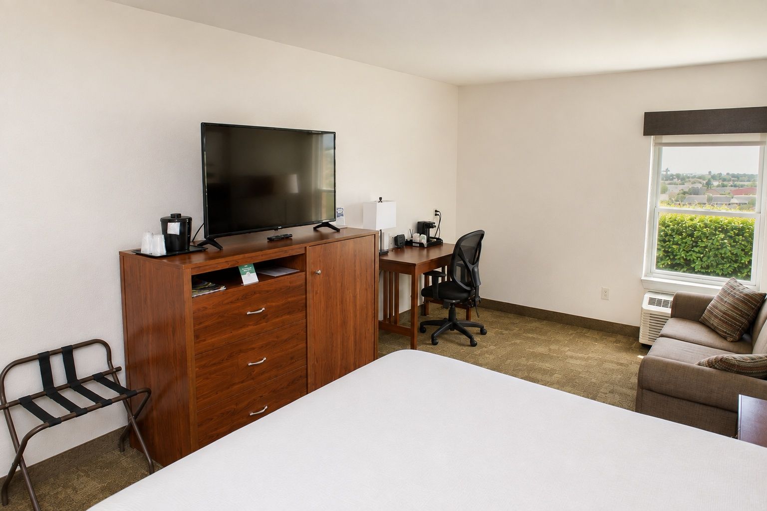 A hotel room with a bed, a desk with a chair, a television on a wooden cabinet, and a window with a view.