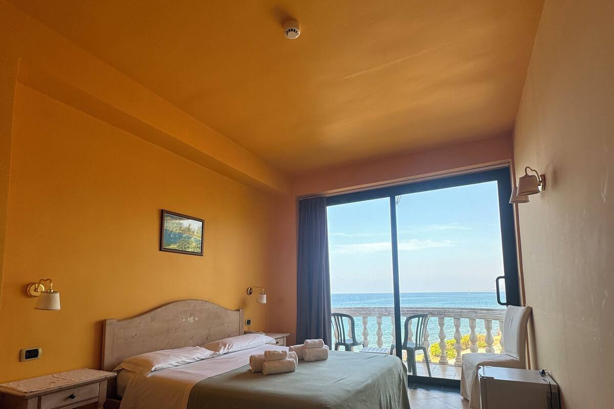 Ein Hotelzimmer mit einem Bett, einem Nachttisch, einer Lampe, einem Sessel, einem kleinen Tisch und Meerblick durch ein Fenster.
