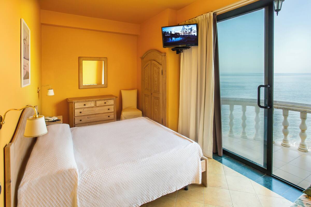 Ein Hotelzimmer mit Bett, Schminktisch, Fernseher und einem Balkon mit Meerblick.