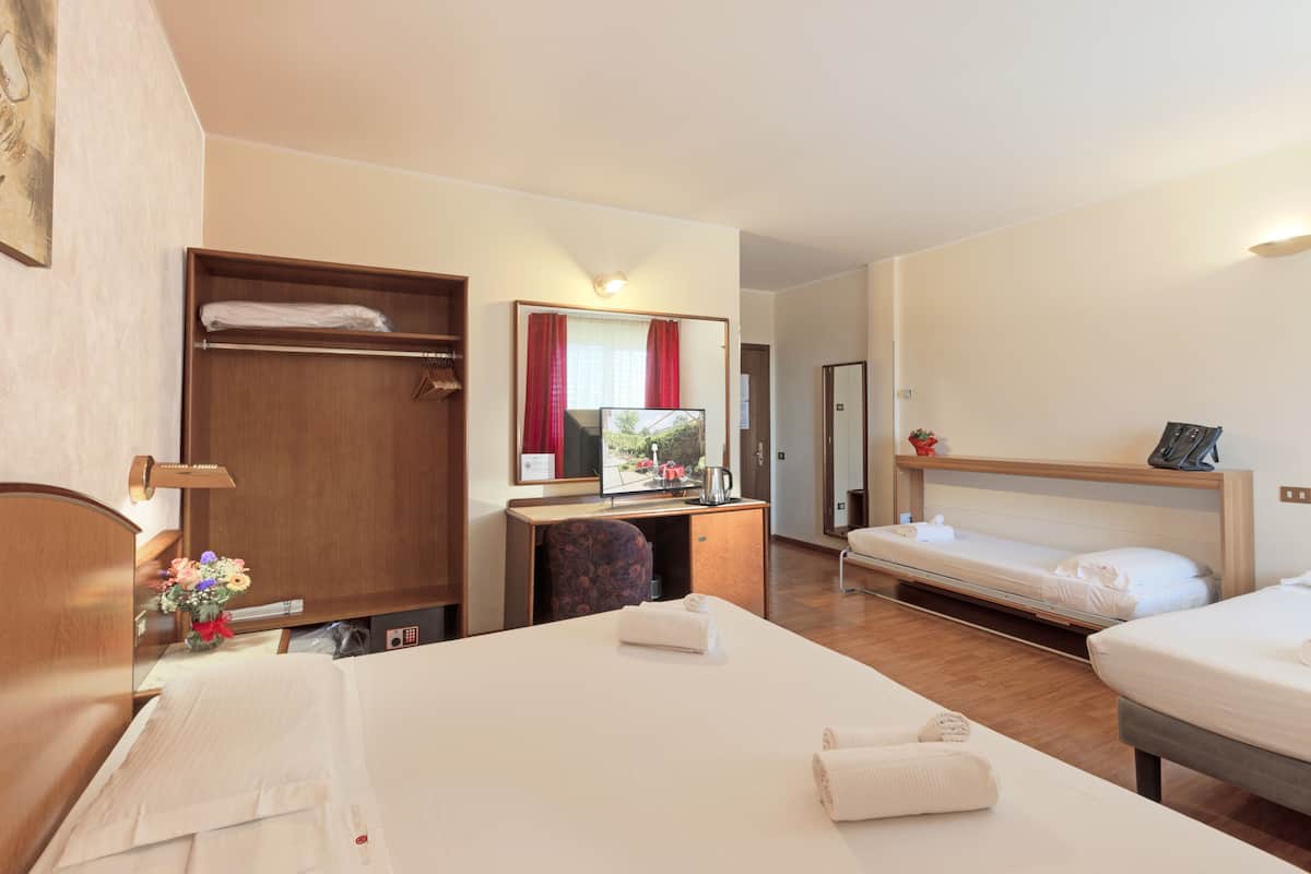 A hotel room with a bed, a desk with a mirror, a wardrobe, and a small table with a vase of flowers.