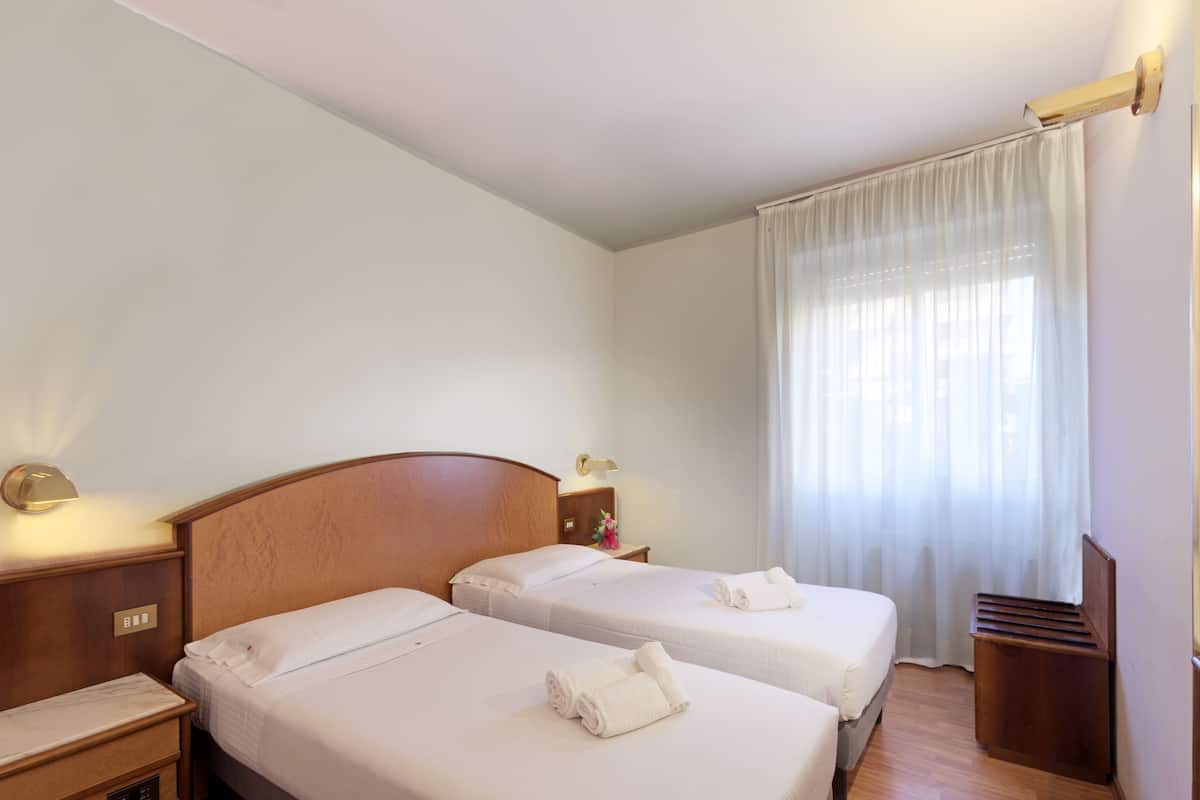 A hotel room with two beds, a wooden headboard, bedside lamps, a nightstand, and a window with sheer curtains.