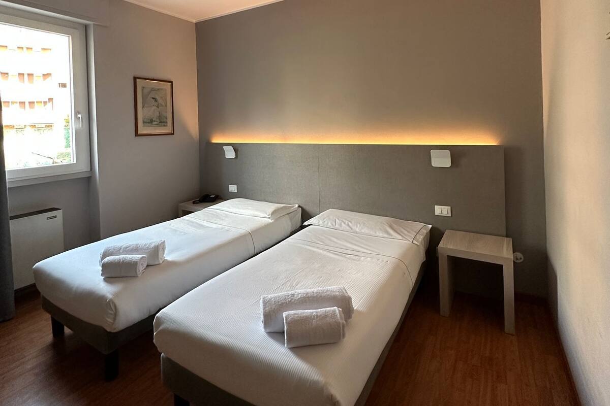 A room with two beds, a small bedside table, a window, and a wall-mounted light fixture.