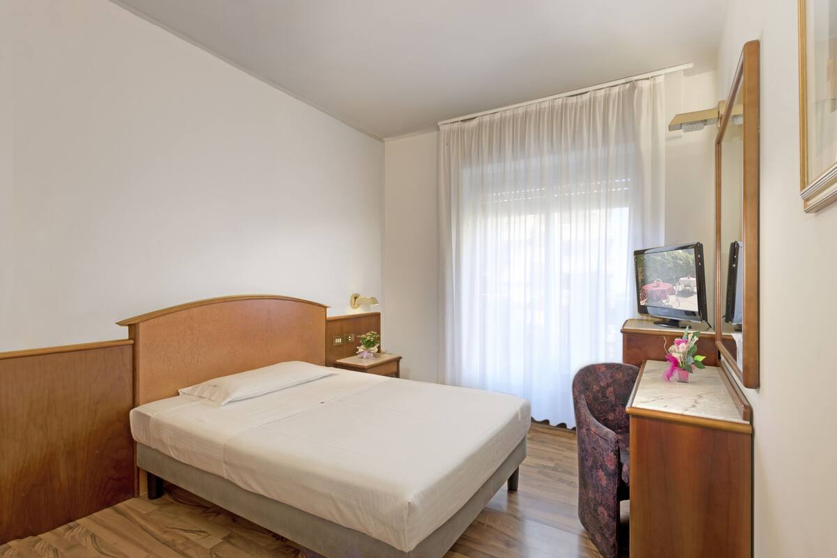A hotel room with a bed, a desk with a mirror, a chair, a television, and a window with curtains.