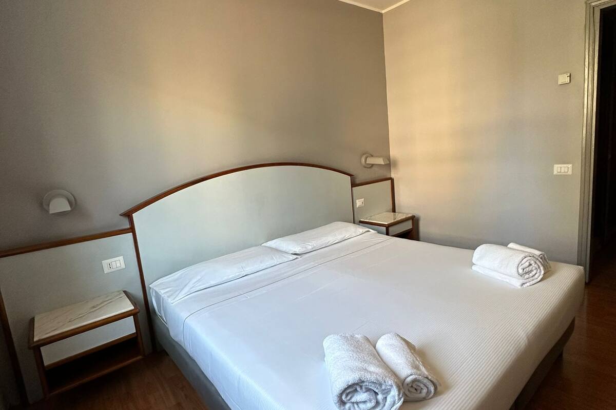A neatly made bed with white linens and two rolled towels, a wooden nightstand with a lamp, and a wooden floor.