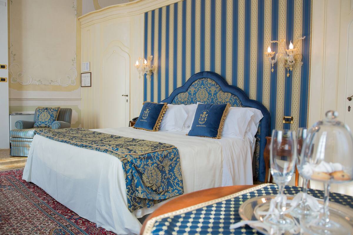 A hotel room with a large bed, a small table with glasses, and a patterned rug.