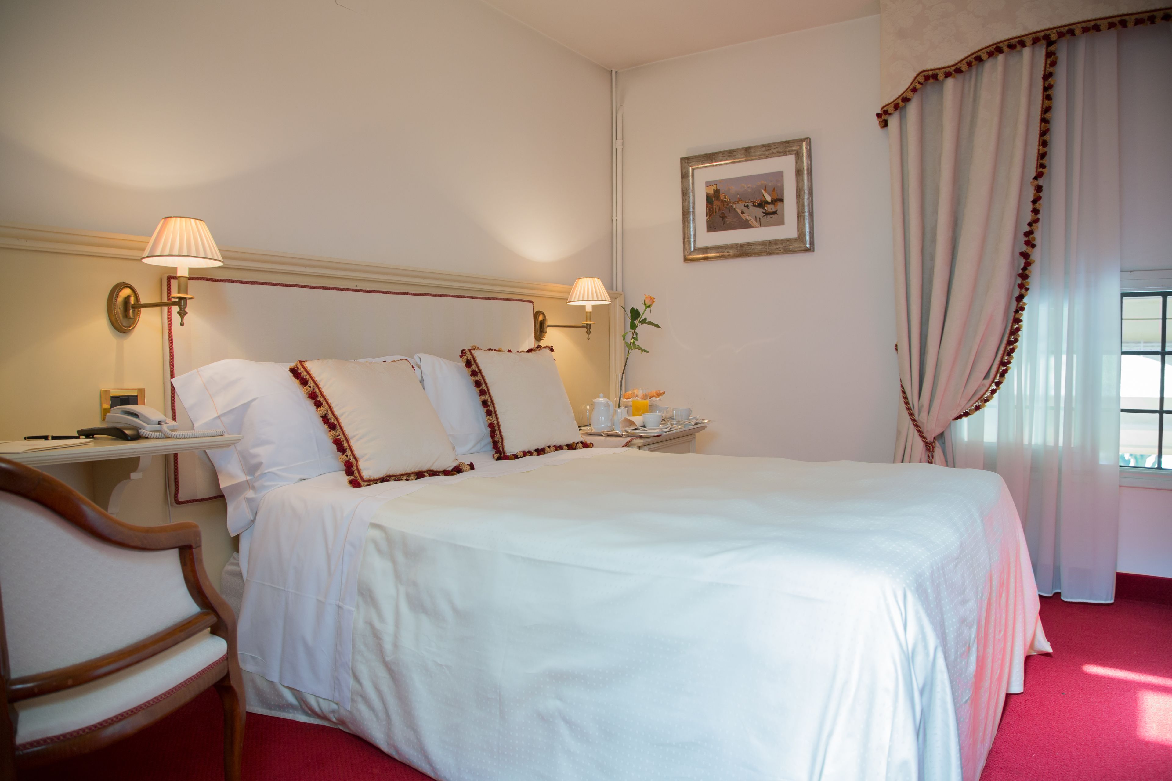 Un dormitorio de hotel con una cama grande, dos mesitas de noche, una silla, una lámpara, un cuadro en la pared y una ventana con cortinas.