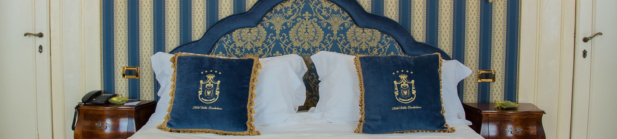 A bed with a blue headboard and two pillows with a crest emblem, flanked by two wooden nightstands with a telephone on one.