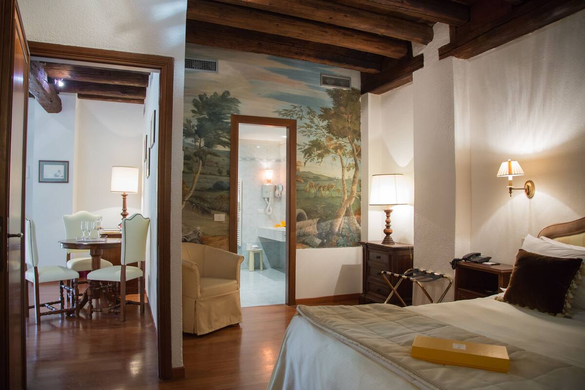 A hotel room with a bed, a desk with a lamp, a chair, and a dining table with chairs.