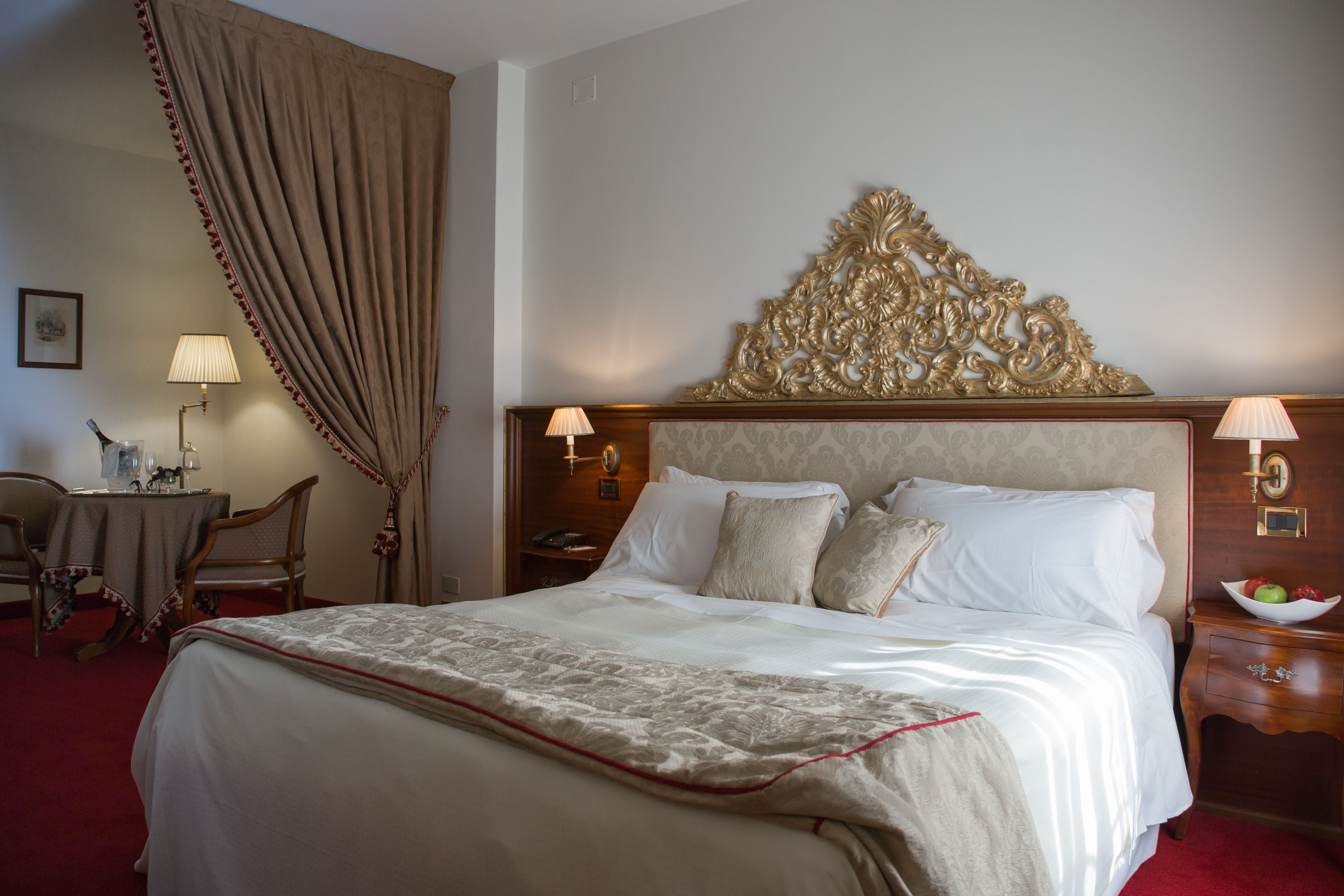 Quarto de hotel com uma cama grande, cabeceira de madeira, duas mesas de cabeceira com abajures, uma escrivaninha com cadeira e uma janela ampla com cortinas.