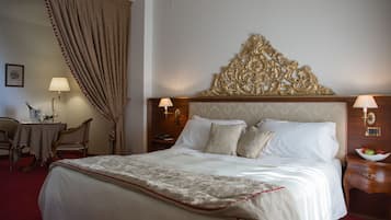 Quarto de hotel com uma cama grande, cabeceira de madeira, duas mesas de cabeceira com abajures, uma escrivaninha com cadeira e uma janela ampla com cortinas.