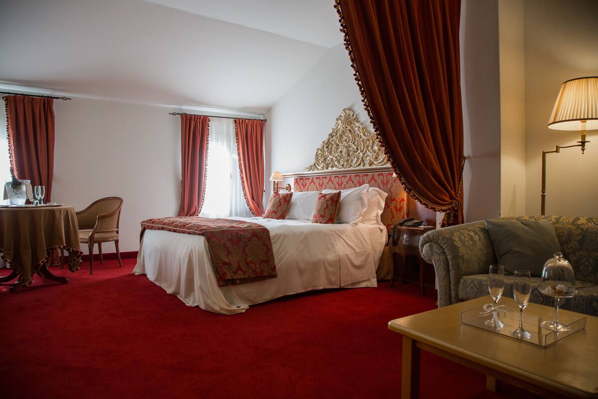 A hotel room with a large bed, a sofa, a table with wine glasses, and a lamp.