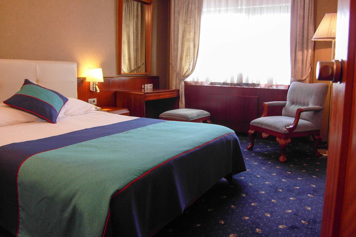 Habitación de hotel con cama, escritorio, silla, lámpara y ventana con cortinas.