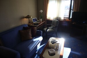 Una habitación de hotel con un sof á azul, un escritorio con una computadora portátil, una silla, una mesa de centro con un control remoto y un televisor.