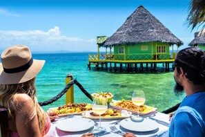 A dining table with plates of food and drinks, overlooking a turquoise sea and a thatched-roof structure. - Punta Caracol Acqua Lodge (Bocas del Toro)