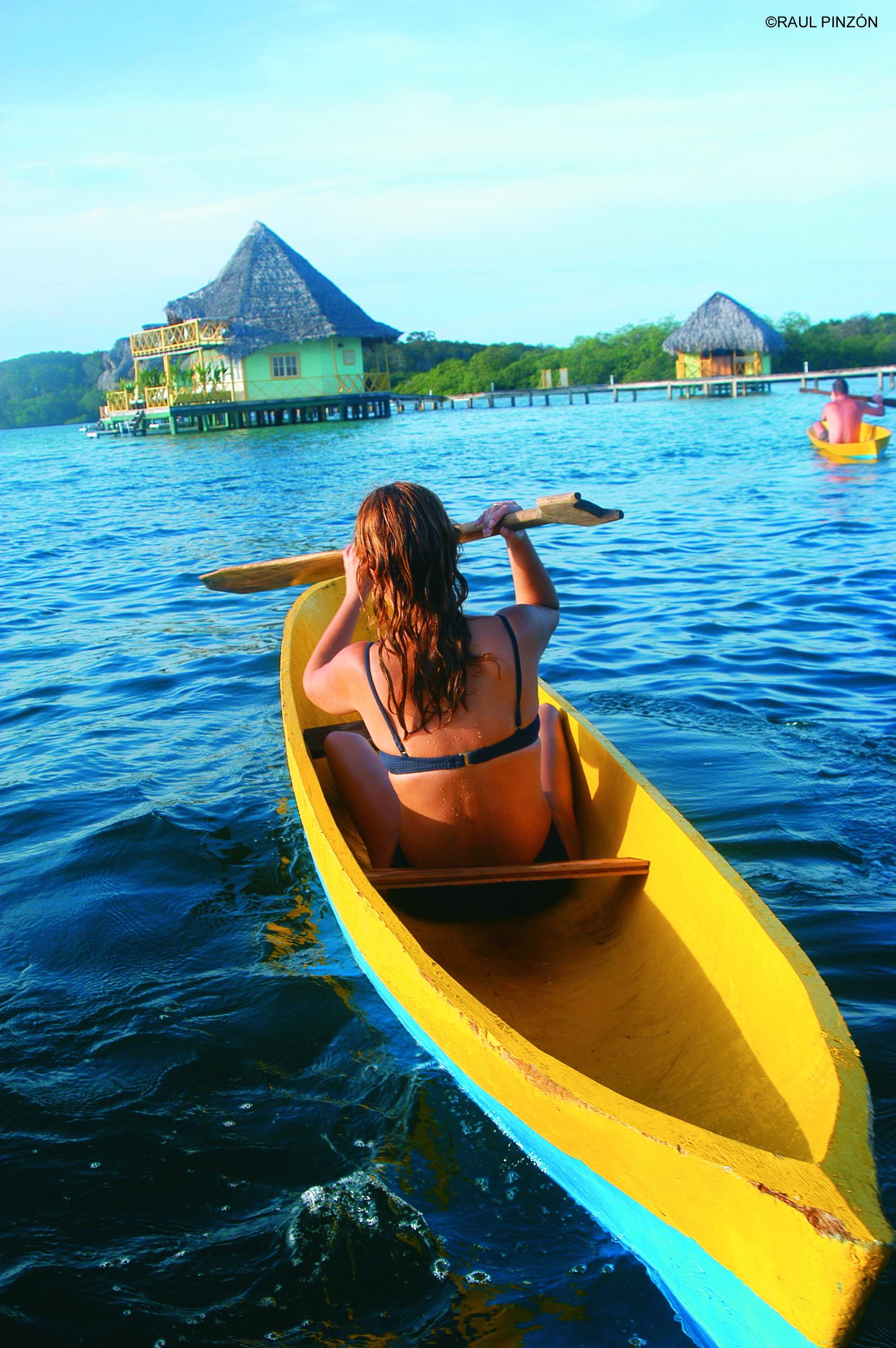 A person in a yellow canoe on calm water, with a house and thatched-roof structures in the background.