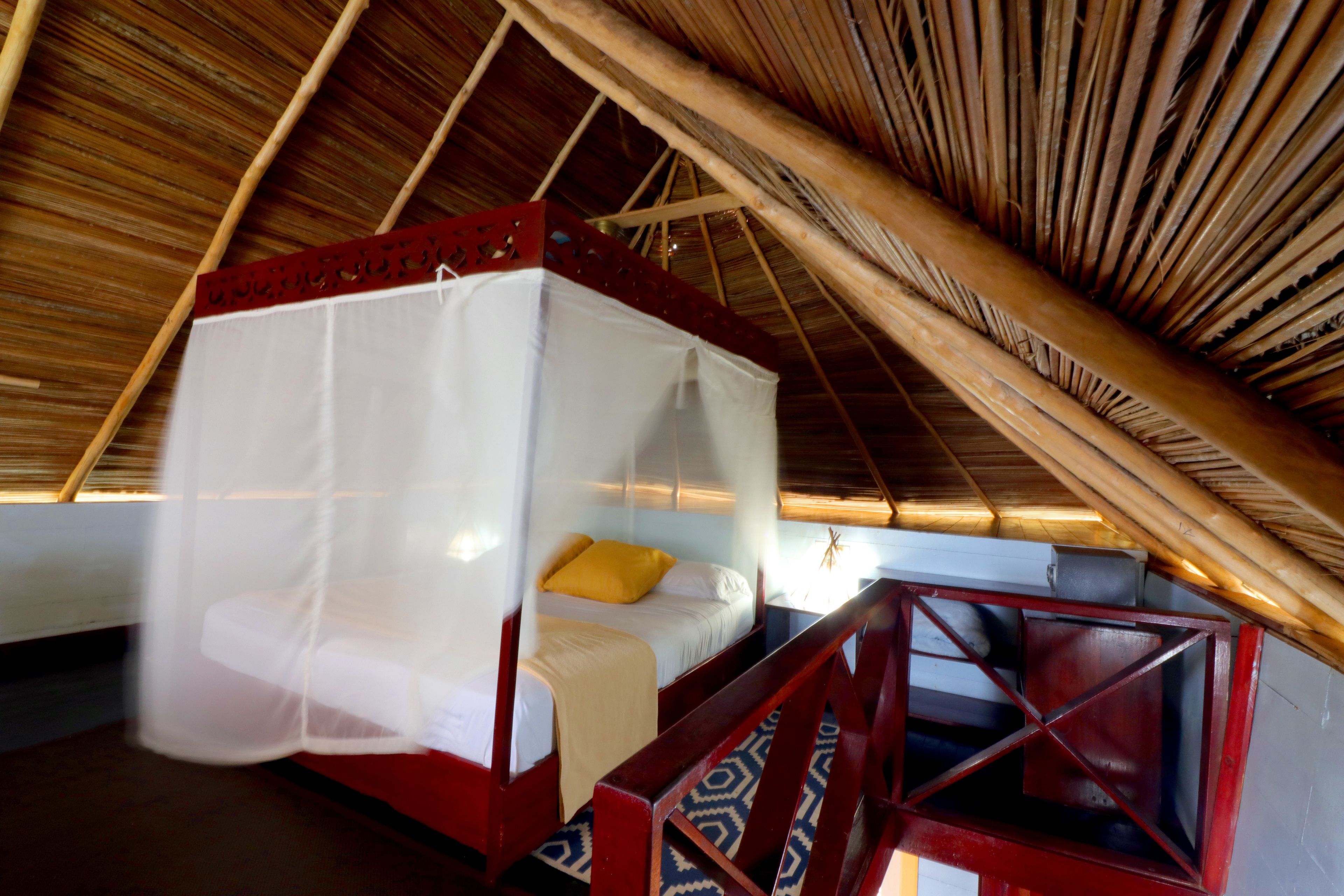 A room with a bed under a thatched roof, a wooden bench, and a small table with a chair.