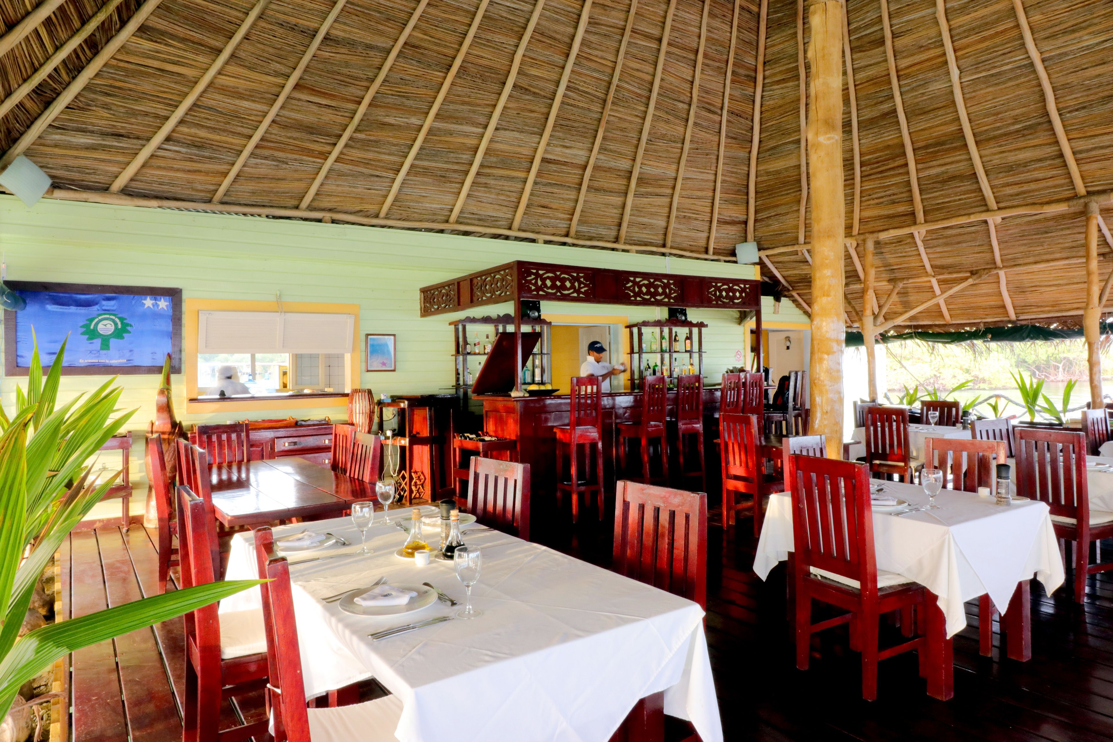 Un restaurant avec des tables et des chaises en bois, un toit de chaume et une vue sur l’extérieur.