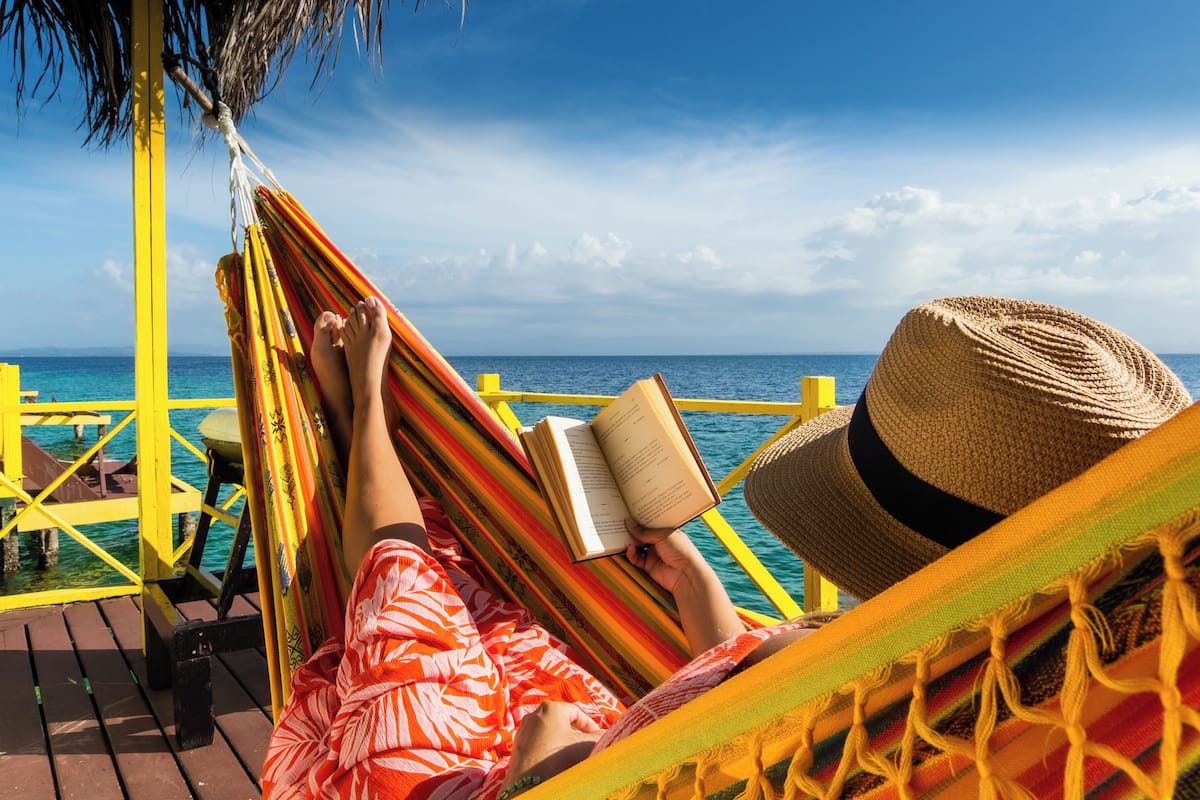 A person relaxing on a hammock with a book, overlooking a clear blue sea from a wooden deck with yellow railings.